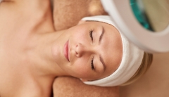 A woman wearing a white cloth headband and her head resting on rolled towel relaxes. Part of a magnifying is seen peaking out in the bottom-right of the picture. 